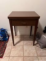 Front view of the vintage sewing table showing single drawer and legs, wood finish, on tiled floor.