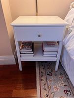 Front view of the white nightstand showing one drawer with silver knob and open bottom shelf with books, placed next to a bed on a wooden floor with a patterned rug.