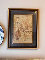 Full view of framed wood carved art, showing village huts, a tree, a fence, and a single figure. Good lighting and image captures frame and carved details.