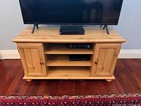 Front view of wooden TV stand with flat top, two side cabinets with wooden knobs, and two central open shelves. Floor and wall visible in background.