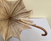 View of the open parasol showing curved wooden handle, lace trimmed fabric canopy with discoloration and fraying, visible metal ribs supporting canopy.