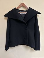 Front view of black wool jacket on wooden hanger against beige wall