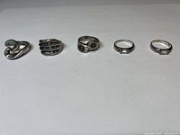 Front view of five sterling silver rings arranged in a row showing different sizes and styles including a modern abstract design, hand motif, stone set rings.