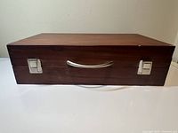 Closed wooden carrying case, dark brown with metal latches and central handle, for storage and transport of art supplies.