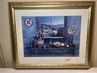 Framed print showcasing a bright still life scene with flowers, fruit, and candles on a rustic blue table, visible through a window revealing a Mediterranean village.