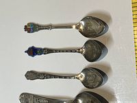 Photo showing top view of 7 souvenir spoons with enamel decorating the handles on two spoons and engraved names on bowls.