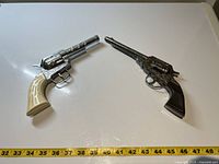 Two vintage revolver cap gun toys side by side on a flat white surface, showing scale with measuring tape indicating length around 11 to 12 inches each.