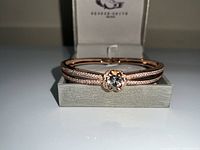 Front angle view of rose gold plated women's bracelet showcasing the large central cubic zirconia stone set in a flower-like setting with two rows of smaller stones on the band, placed in light gray George Smith branded jewelry box.