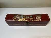 Front view of a deep red pillow-shaped Chinese wooden jewelry box with gold and pastel floral designs and a metal latch.