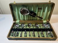 The full set displayed inside original emerald-green lined wooden case showing one large serving spoon resting in lid and six individual sets of spoon and fork held in black velvet straps.