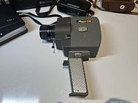 Side view of vintage gray Lumicon Zoom Auto-Eye 8mm camera with pistol grip handle and black zoom lens on white surface.