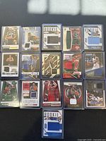 Top-down view of 15 basketball rookie trading cards each encased in protective hard plastic holders. Cards feature embedded jersey fabric patches in various colors and designs, including numbered autograph card.