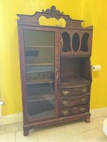Front view of the antique wooden display cabinet showing glass-fronted shelving section beside an open carved wood section with drawers below.