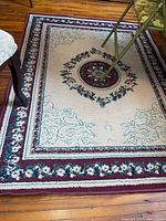 Area rug with burgundy border, cream background with floral medallion pattern, laid on hardwood floor with a chair partially visible.