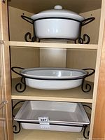 Three white ceramic baking dishes on shelves with black metal holders: top is round with lid, middle is oval, bottom is rectangular dish.