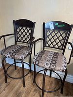 Two metal framed bar chairs with brown lattice backs, upholstered seats in patterned fabric, armrests, and circular foot rails shown from front angle against a beige wall.