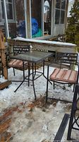 Full view of the bistro set on a snowy deck showing square glass top table and two metal frame chairs with cushions.
