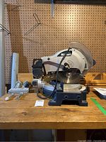 Side and front views showing Mastercraft compound mitre saw with attached blade guard and included clamps on wooden workbench.