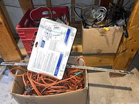 View of cardboard box filled with orange extension cords with two-pack smoke alarms on top and a white meter stick placed across box