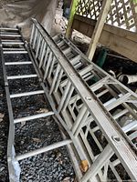 Photo showing the aluminum extension ladder lying on gravel surface under a deck area, showing ladder rungs and side rails in detail.