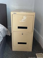 Front view of beige Admiral 2-drawer filing cabinet next to bed, showing drawer handles and worn label/sticker.