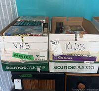 Two cardboard boxes, one labeled VHS and another labeled Kids with assorted kids movies DVDs and VHS in original sleeves and cases.