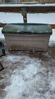 Front view of the plastic patio storage box outdoors on a wooden deck with some snow around it, showing the beige base and green lid with weathering and dirt marks.
