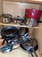 Photo showing pots and pans arranged on a wooden shelf including red enameled pots, metal pans, and glass lids.