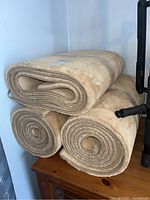 Three beige carpet runners rolled up and stacked on a wooden surface against a white wall.