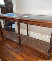 Side view of vintage sofa table showing glass top, turned wooden legs, and cane bottom shelf on wooden floor.