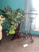 Photo showing two heavy metal bathroom tissue holders next to a faux leafy plant in a dark wooden pedestal stand indoors by a window with blue curtains.