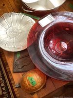 Photo showing stacked glass bowls and plates: clear flower-shaped bowl, matching clear bowl and plate, red decorative bowl. Cast iron wall hanging and amber turtle light partially visible.