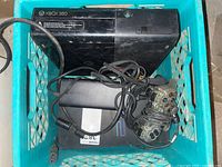 Xbox 360 console model 1538, power brick, cables, and PlayStation 2 console inside a green milk crate.