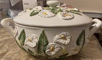 Photo showing side view of cream ceramic pot with 3D white flowers, green leaves, two handles, and lid with knob.