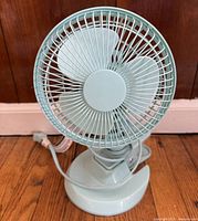 Front view of light blue desk fan showing blades, protective grille, and base with power cord.