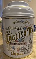 Front view of the white metal tea canister labeled 'Traditional English Tea Fine Breakfast Tea' with floral and gold decoration and quantity indication of 240 tea bags.