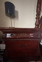 Front view of wooden nightstand showing three drawers with black metal handles and floral carving on top drawer