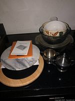 Photo showing silver plate cream and sugar set, stacked ceramic bowls with floral pattern, round marble trivet with wooden base, and a digital food scale with manual on a black countertop.