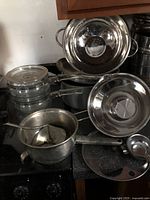 Photo showing all seven items: glass double boiler with glass lid, metal upper pot, metal base pot, metal colander, metal ricer with press, metal boiler pan with long handle, and egg poacher insert.
