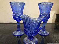 Two blue glass goblets with silhouettes and one pitcher with Monticello design arranged on a surface.