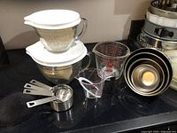 Photo showing the set of Pampered Chef glass mixing bowl with lid, clear plastic measuring cup, metal measuring cups and metal mixing bowls on kitchen counter.