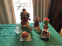 Overall view of the collection including wind-up musical Santa, glass Santa figurine, various smaller Santa figurines, Grinch in Santa attire, a Christmas ball ornament, and a small ceramic house decoration with painted Santa faces.