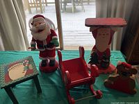 Photo showing all four Christmas-themed items: battery-operated Santa figure, small Santa face footstool, Santa-themed table, and red wooden sleigh.