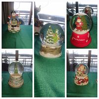 Five Christmas-themed snowglobes with festive characters and winter scenes on green table covering near window with lake view in background.