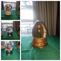 Four Christmas-themed snow globes displayed on green fabric near a window with outdoor lake view.