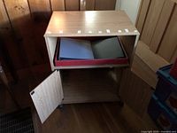 Wooden computer cabinet with two doors open showing an inner shelf and a tray containing folders. The tabletop shows damage or wear.