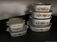Seven CorningWare casserole dishes arranged in two stacks showing vegetable designs and lids of plastic and glass types.
