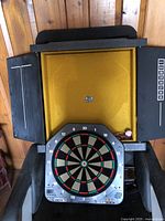 Electronic dartboard mounted inside wooden cabinet with doors open showing dartboard face and some darts and chalk inside.
