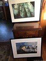 Photo showing two framed nature prints leaning against a piece of furniture. One print depicts bamboo leaves, the other shows mountain peaks.