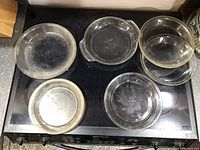 Five assorted clear glass pie plates arranged on a black surface. Includes one pie plate with two small handles. Plates vary between approximately 6 to 8 inches diameter.